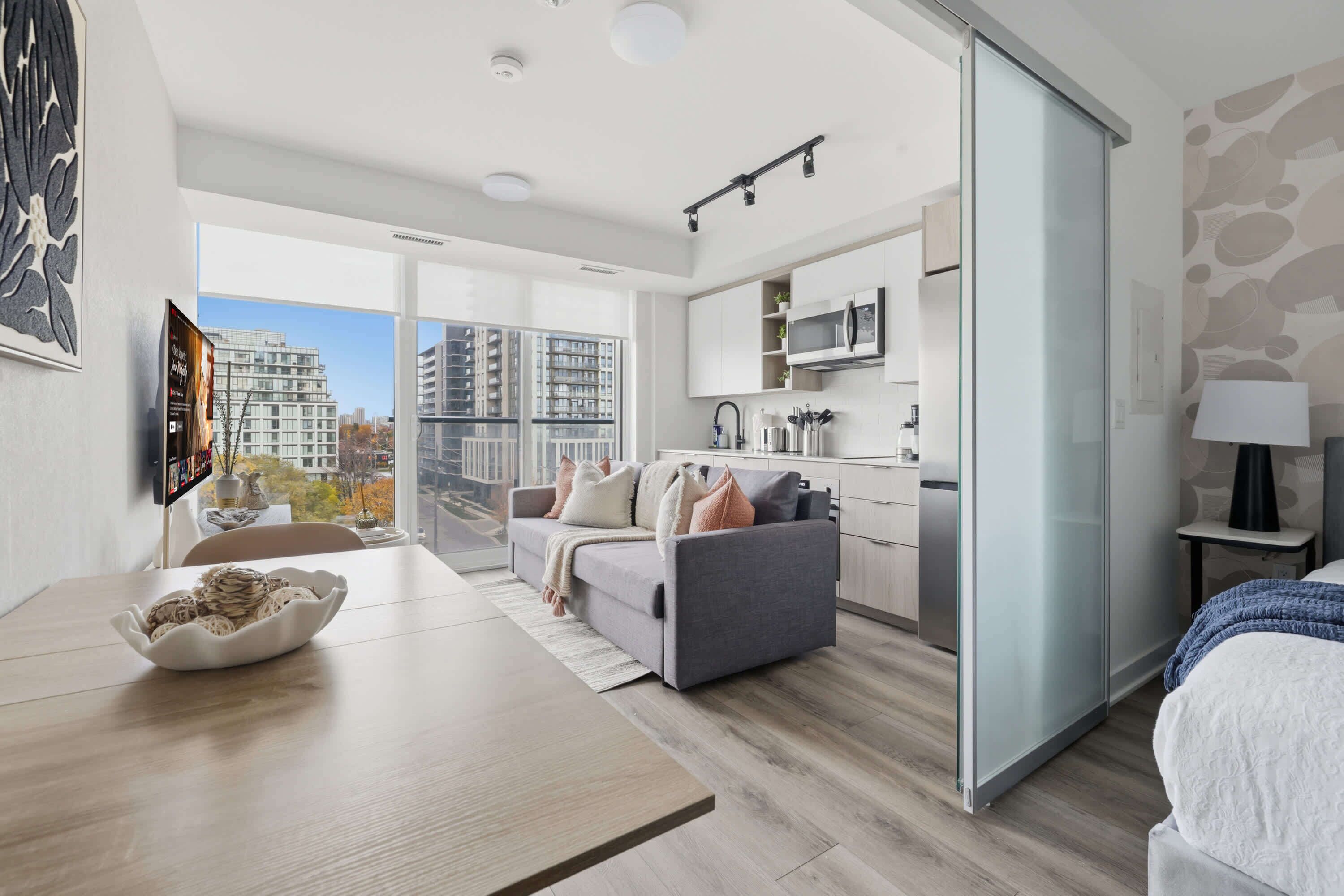 A modern apartment with a dining area, a sofa, a kitchen, and a large window offering a city view.