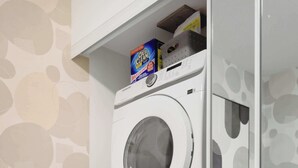 A laundry area with two stacked washing machines, a mirror, a small table with a lamp, and a bed with pillows.