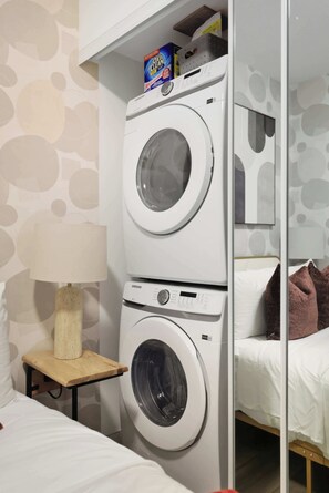 Un espace buanderie comprenant deux machines à laver superposées, un miroir, une petite table avec une lampe et un lit avec des oreillers.