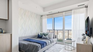 A modern bedroom with a large bed, a balcony with a city view, a small desk, and a TV.