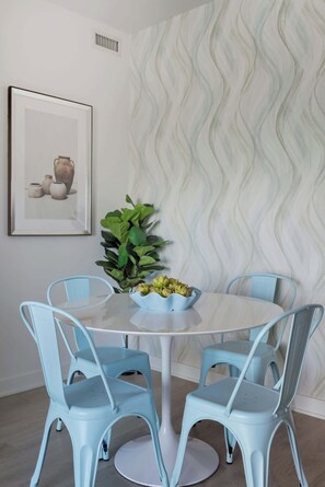 Uma sala de jantar com uma mesa redonda, quatro cadeiras azuis, um vaso com planta e um quadro na parede.
