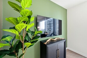 A room with a flat-screen TV mounted on a wall, a dark wooden cabinet below, and a large green plant to the left. - Downtown 1BR Urban Comfort Steps from it All (Toronto)