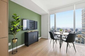 A modern living room with a flat-screen TV, a dark cabinet, a potted plant, a dining table with chairs, and a large window offering a city view. - Downtown 1BR Urban Comfort Steps from it All (Toronto)