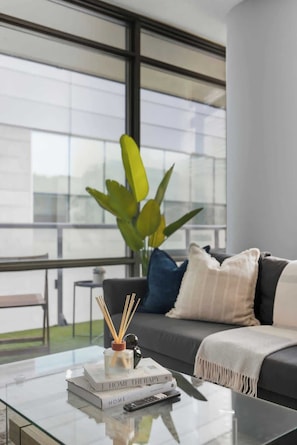 A modern living room with a glass coffee table, a sofa with cushions, a large window, and a potted plant. - Designer Skyline Retreat 2BR w Skyline Terrace! (Toronto)