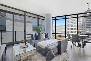 A modern bedroom with a large bed, a glass coffee table, a dining area with a table and chairs, and a view of a cityscape through large windows.