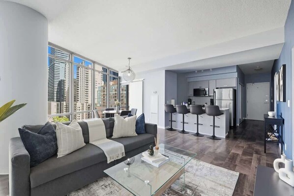 A modern living room with a sofa, coffee table, and a view of the cityscape through large windows.
