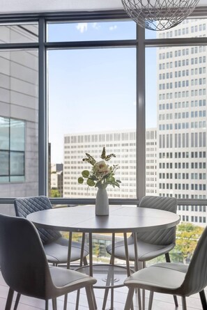 A modern dining area with a round table and six chairs, a vase with flowers, and a view of tall buildings through large windows.