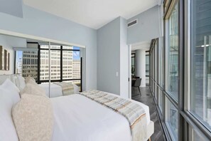 A modern hotel room with a large bed, a mirror, and a view of a cityscape through large windows.