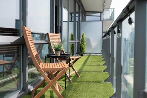 A modern outdoor balcony with a small artificial grass area, two wooden chairs, a small table with a potted plant, and a view of the cityscape through large windows. - Modern 2BR Haven, Luxury Stay in Financial Dist (Toronto)