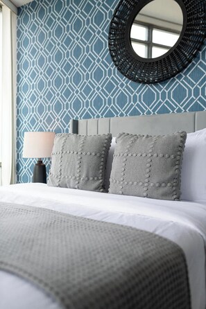 A neatly made bed with a white duvet and grey pillows, a round mirror on the wall, and a patterned blue wallpaper. - Modern 2BR Haven, Luxury Stay in Financial Dist (Toronto)