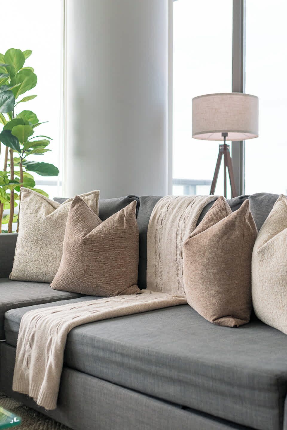 A grey sofa with beige cushions and a throw blanket, positioned near a window with a lamp.