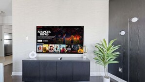 Moderni olohuone, jossa on litteänäyttöinen televisio, jossa näkyy Netflixin käyttöliittymä, tumma puulattia, valkoinen konsolipöytä ja ruukussa oleva viherkasvi.