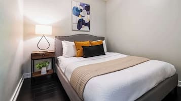 Een net opgemaakt bed met hoofdeinde, nachtlampje, een klein plantje en een kunstwerk aan de muur.