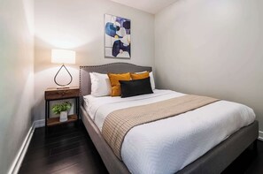 A neatly made bed with a headboard, a bedside lamp, a small plant, and a wall art piece. - Elevated Downtown 3BR w Views & Free Parking (Toronto)