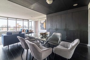 A modern living room with a glass dining table, grey upholstered chairs, a dark-colored sofa, and a large window offering a city view. - Elevated Downtown 3BR w Views & Free Parking (Toronto)