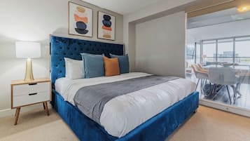 Une chambre moderne avec une tête de lit capitonnée, un grand lit avec de nombreux oreillers, une table de chevet avec une lampe et une vue sur un coin repas à travers une porte vitrée.
