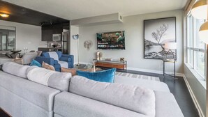 A modern living room with a large grey sofa, a dining area with a glass table, and a kitchen in the background. - Elevated Downtown Living Luxe 3BR w Views! (Toronto)