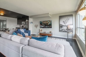 A modern living room with a large grey sofa, a dining area with a glass table, and a kitchen in the background. - Elevated Downtown Living Luxe 3BR w Views! (Toronto)