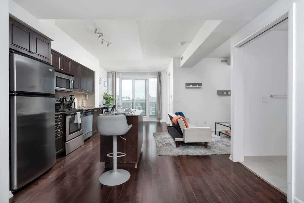 A modern kitchen with stainless steel appliances, a dining area with a bar stool, and a living space with a sofa and a view of the city.