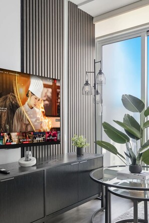 Ein modernes Wohnzimmer mit einem großen Flachbildfernseher an einer Wand mit vertikaler Wandverkleidung, einem Glastisch mit einem Buch und einem Topfpflanzen.