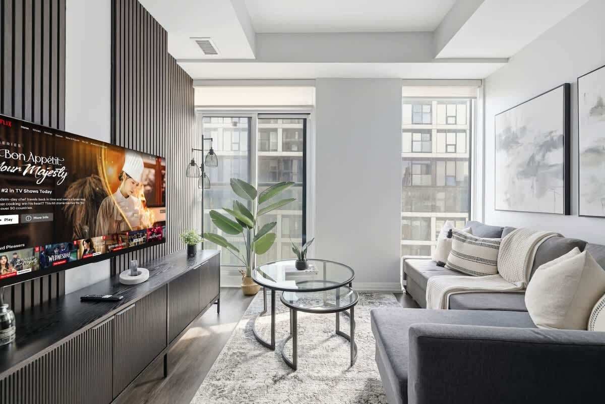 A modern living room with a flat-screen TV, a glass coffee table, a grey sofa, and a large window.