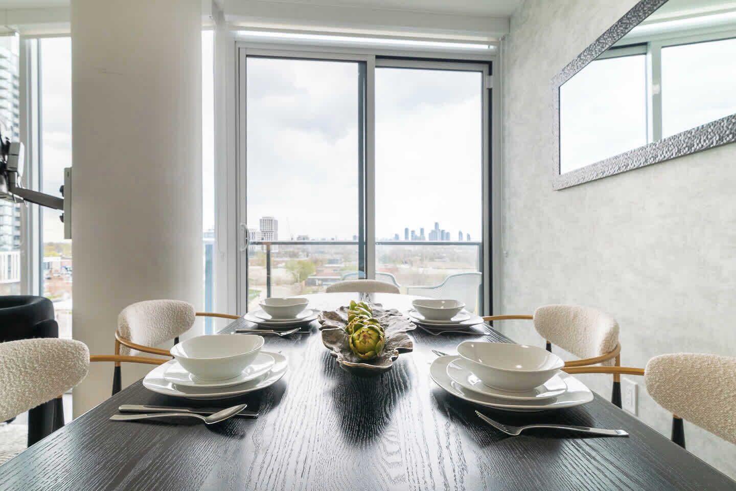 A dining table set for six with white plates and bowls, a centerpiece of artichokes, and a city view through large windows.