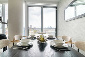 A dining table set for six with white plates and bowls, a centerpiece of artichokes, and a city view through large windows.