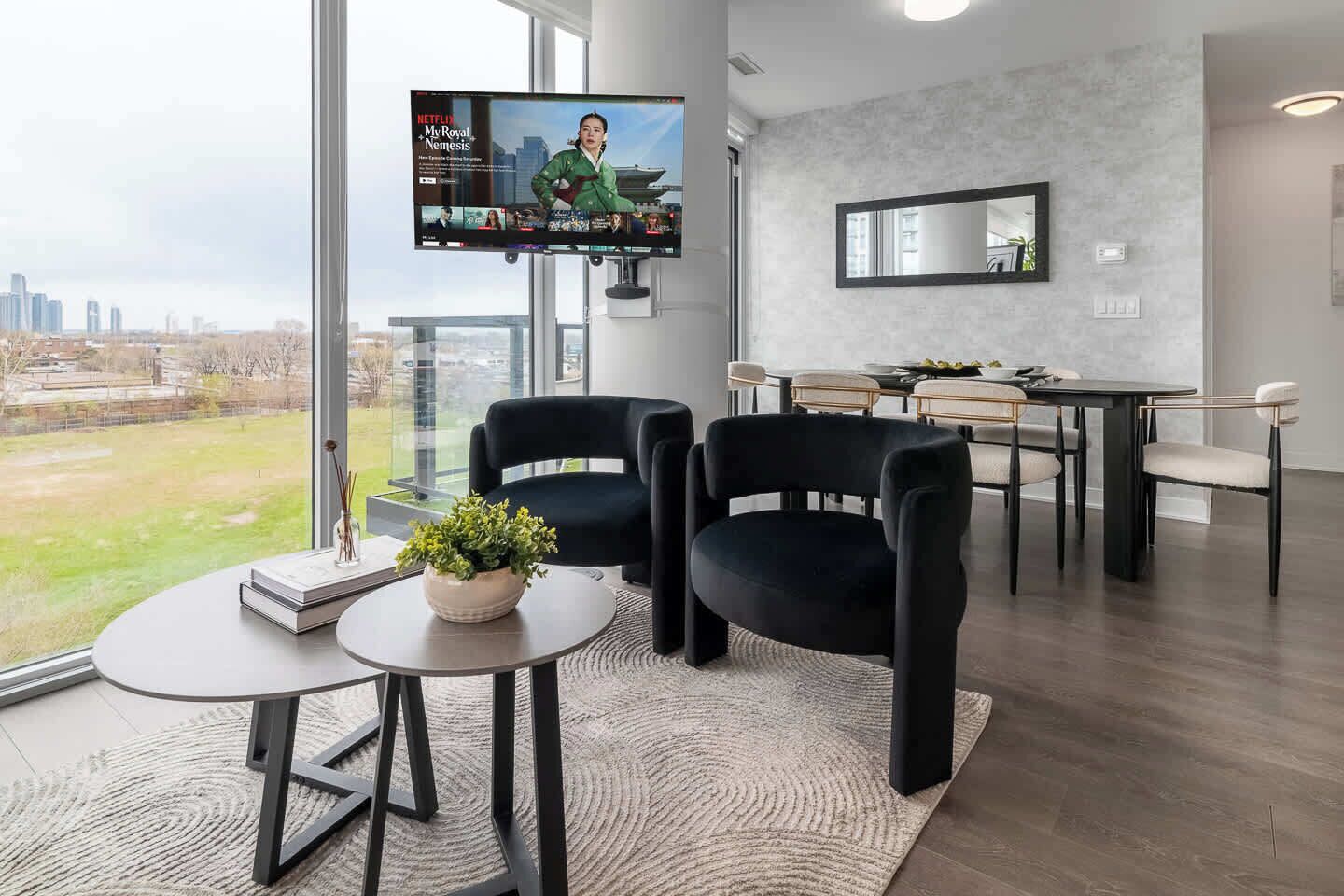 A modern living room with a large TV, black chairs, a small table with a plant, and a city view.