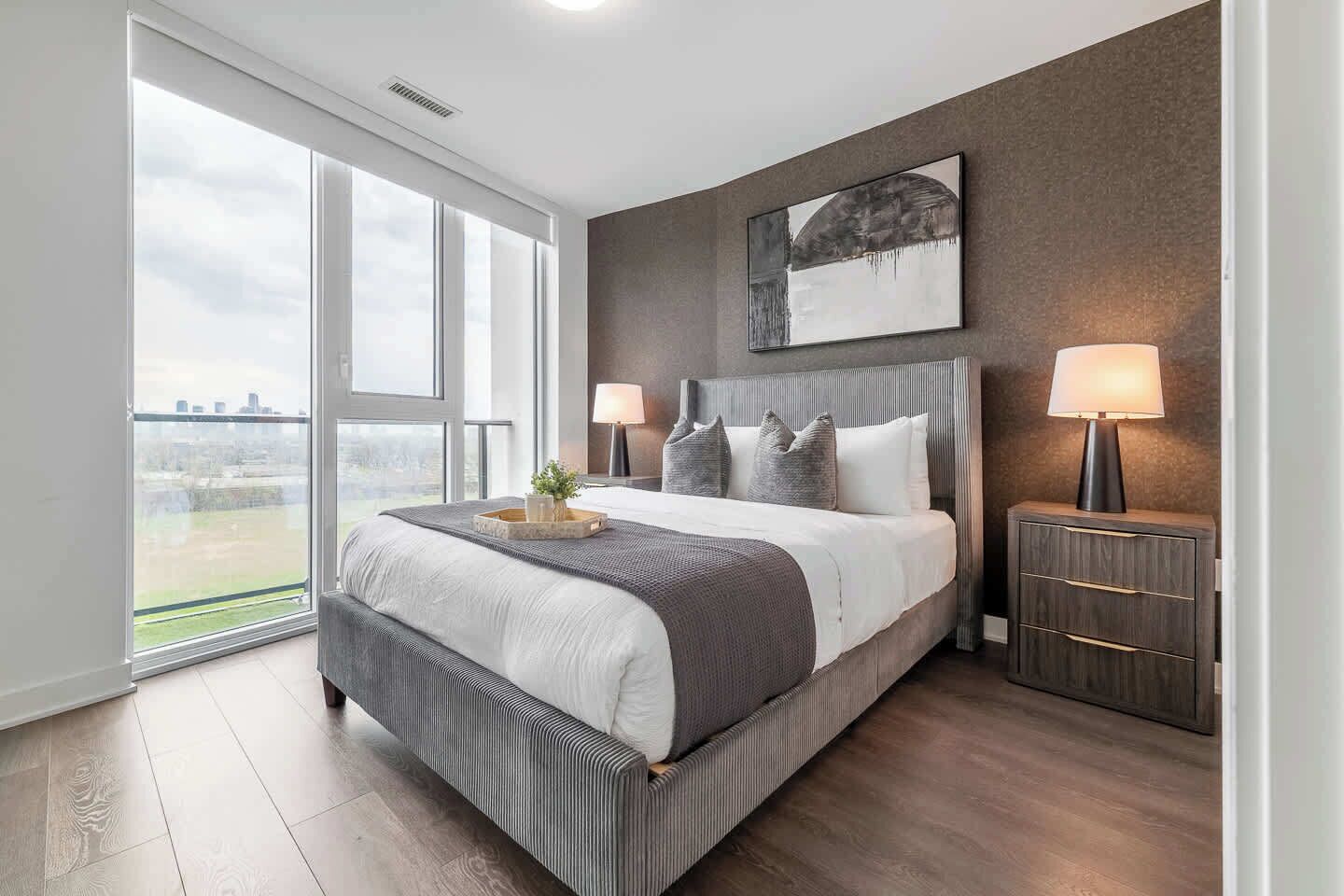 A modern bedroom with a large bed, two bedside lamps, a view through a sliding door, and a cityscape visible outside.