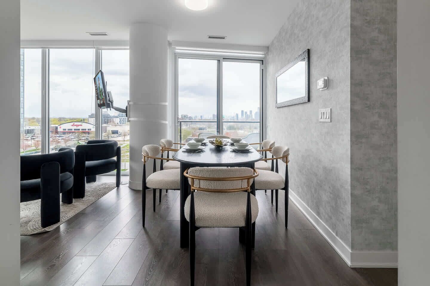 A dining area with a round table, six chairs, and a city view through large windows.