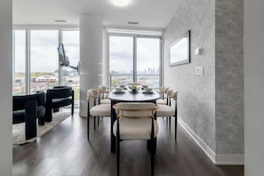 A dining area with a round table, six chairs, and a city view through large windows.