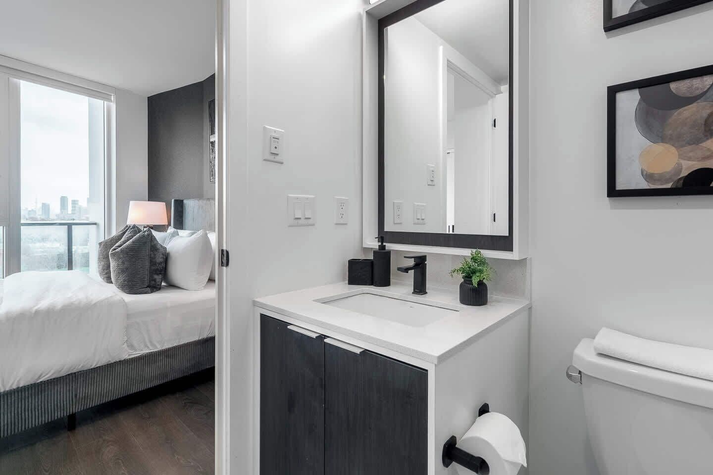 A modern hotel room with a large bed, a bedside table with a lamp, a bathroom with a sink, mirror, and toilet.