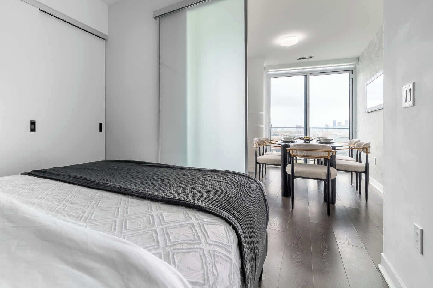 A modern hotel room with a large bed, a dining area with a table and chairs, and a view through a large window.