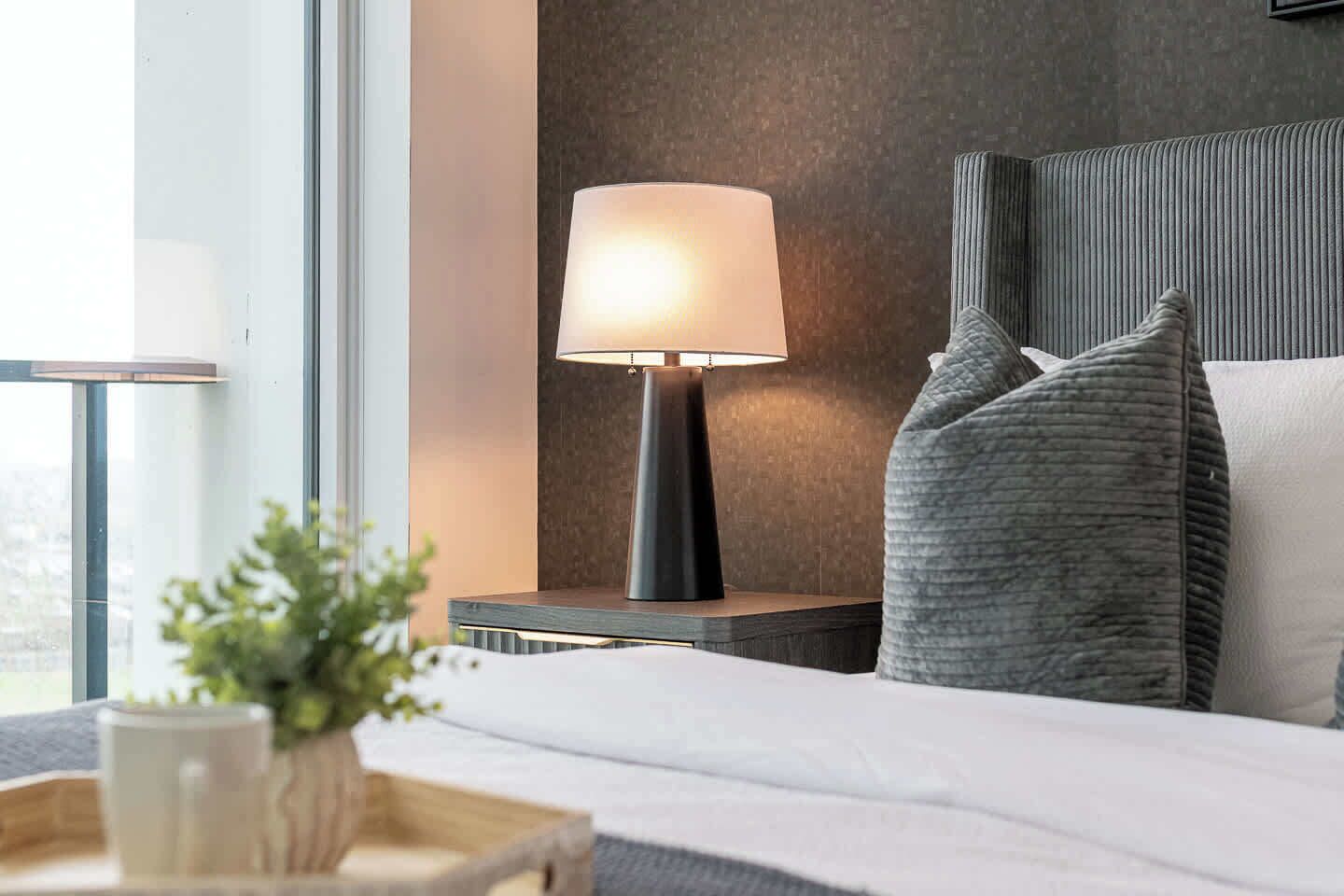 A hotel room with a bed, a bedside table with a lamp, a window, and a view of the city.
