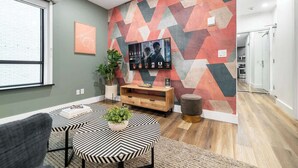 Ein modernes Wohnzimmer mit Wandpaneelen in geometrischem Muster, einem Flachbildfernseher, einem Holzkonsolentisch und einem Essbereich mit gestreiftem Tischlein.