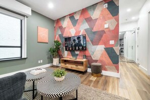 Ein modernes Wohnzimmer mit Wandpaneelen in geometrischem Muster, einem Flachbildfernseher, einem Holzkonsolentisch und einem Essbereich mit gestreiftem Tischlein.