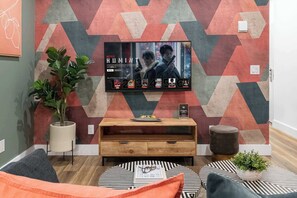 Ett vardagsrum med en platt-TV som är monterad på en vägg med ett geometriskt mönster, ett soffbord i trä, en krukväxt och en soffa.