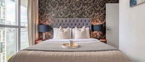 A modern bedroom with a tufted headboard, a large bed, two bedside lamps, and a view of the city through a large window.