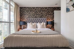 Une chambre moderne avec une tête de lit capitonnée, un grand lit, deux lampes de chevet et une vue sur la ville par une grande fenêtre.