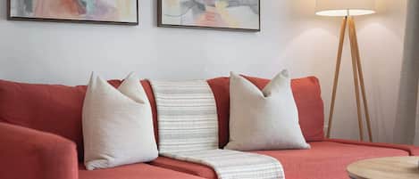 Un sofá rojo con tres almohadones, una manta blanca y una lámpara de pie con base de madera.