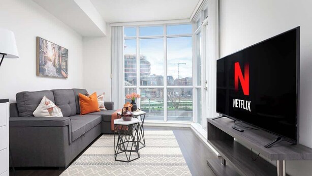 A modern living room with a grey sofa, a coffee table, and a large flat-screen TV displaying the Netflix logo. - Downtown Escape Stylish Condo with City Views (Toronto)