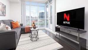 A modern living room with a large flat-screen TV displaying the Netflix logo, a grey sofa with orange cushions, a small round table with a vase, and a patterned rug. - Downtown Escape Stylish Condo with City Views (Toronto)