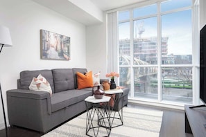 A modern living room with a grey sofa, a coffee table, and a large window offering a view of a cityscape. - Urban Waterfront 1BR | Sleeps 5 (Toronto)