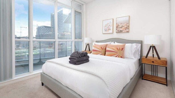 A hotel room with a large window, a bed with white bedding, two bedside lamps, and a small wooden nightstand.