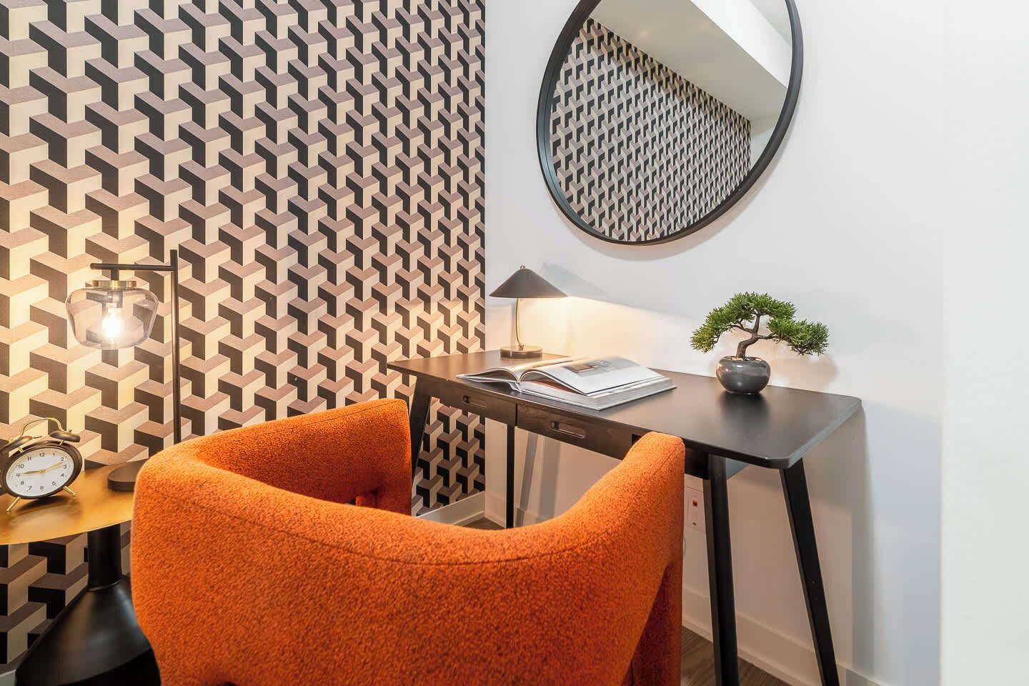 A modern room with a geometric patterned wall, a round mirror, a desk with a lamp, a bonsai tree, and an orange chair.