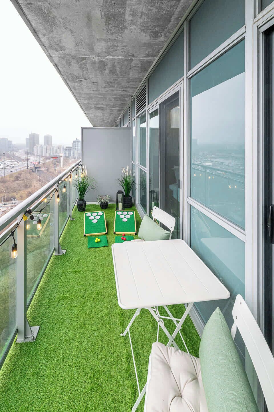 Een balkon met kunstgras, een witte tafel en twee stoelen. Er staan potplanten en er is uitzicht op een stadsgezicht door grote ramen.