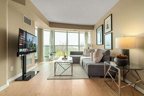 A modern living room with a flat-screen TV, a grey sofa, a glass coffee table, and a lamp.