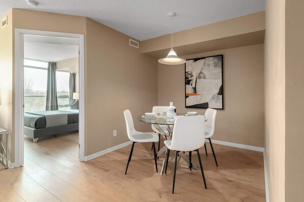 A modern dining area with a glass table and white chairs, a ceiling light, and a painting on the wall.
