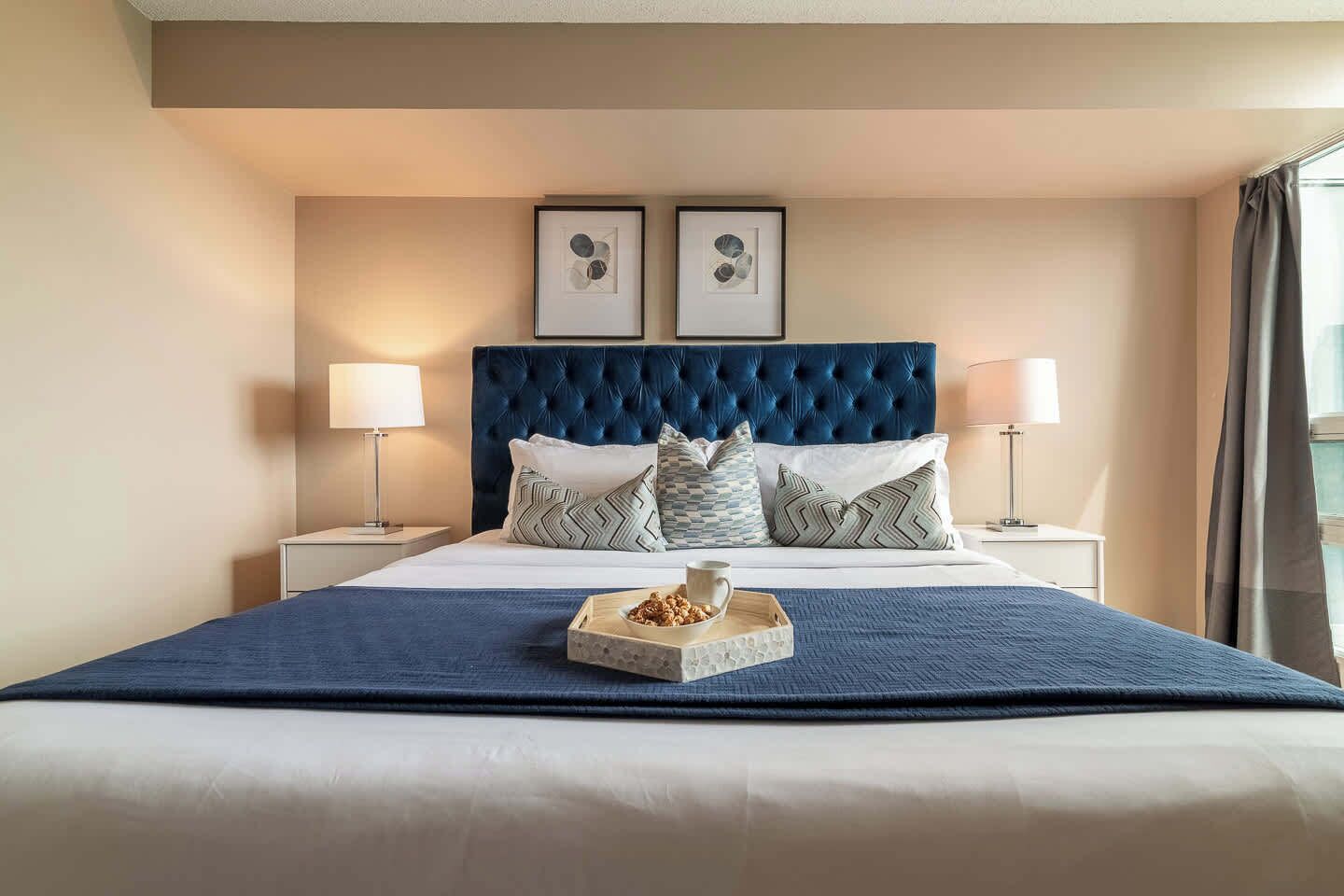 Une chambre d’hôtel avec un grand lit, deux tables de chevet avec des lampes, une tête de lit à motifs capitonnés et deux tableaux encadrés au mur.