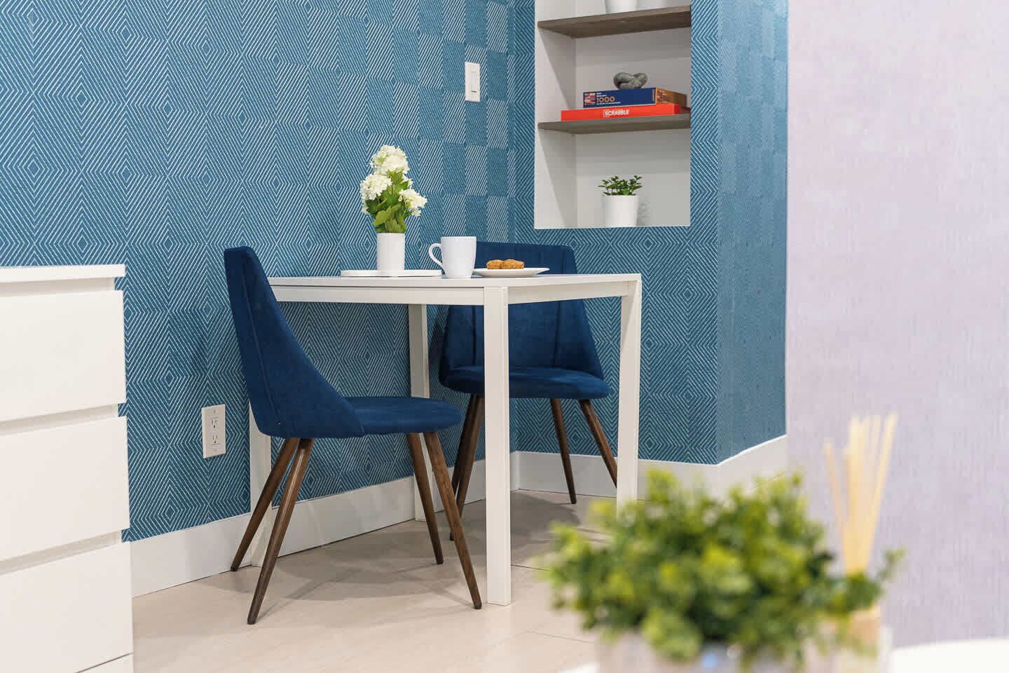 A dining area with a white table and two blue chairs, a vase with flowers, and a shelf with books and a plant.